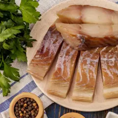 Wędzony czarniak, podobny do dorsza, pokrojony na kawałki, leży na talerzu obok świeżej natki pietruszki i pieprzu.