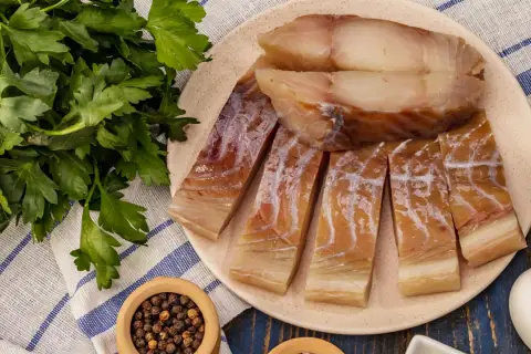 Wędzony czarniak, podobny do dorsza, pokrojony na kawałki, leży na talerzu obok świeżej natki pietruszki i pieprzu.