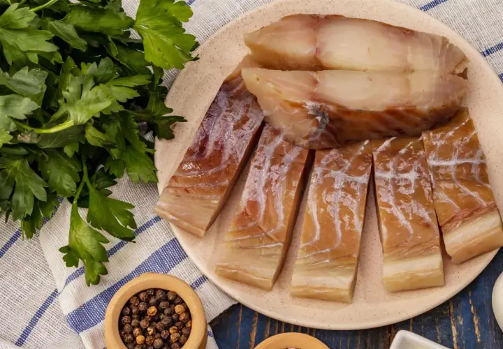 Wędzony czarniak, podobny do dorsza, pokrojony na kawałki, leży na talerzu obok świeżej natki pietruszki i pieprzu.
