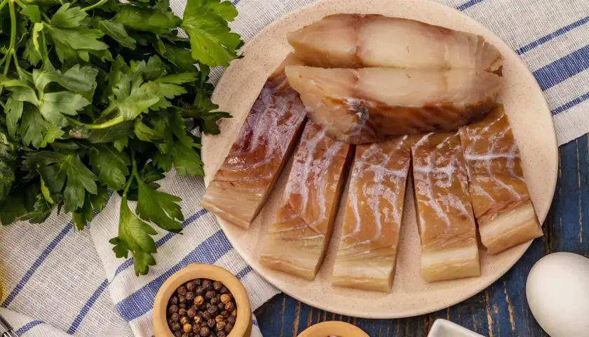 Wędzony czarniak, podobny do dorsza, pokrojony na kawałki, leży na talerzu obok świeżej natki pietruszki i pieprzu.